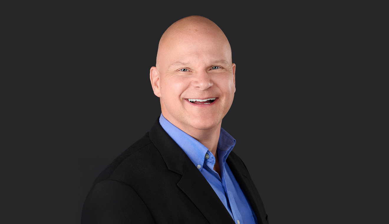 Bald man in a black blazer and blue shirt smiling in a corporate studio headshot